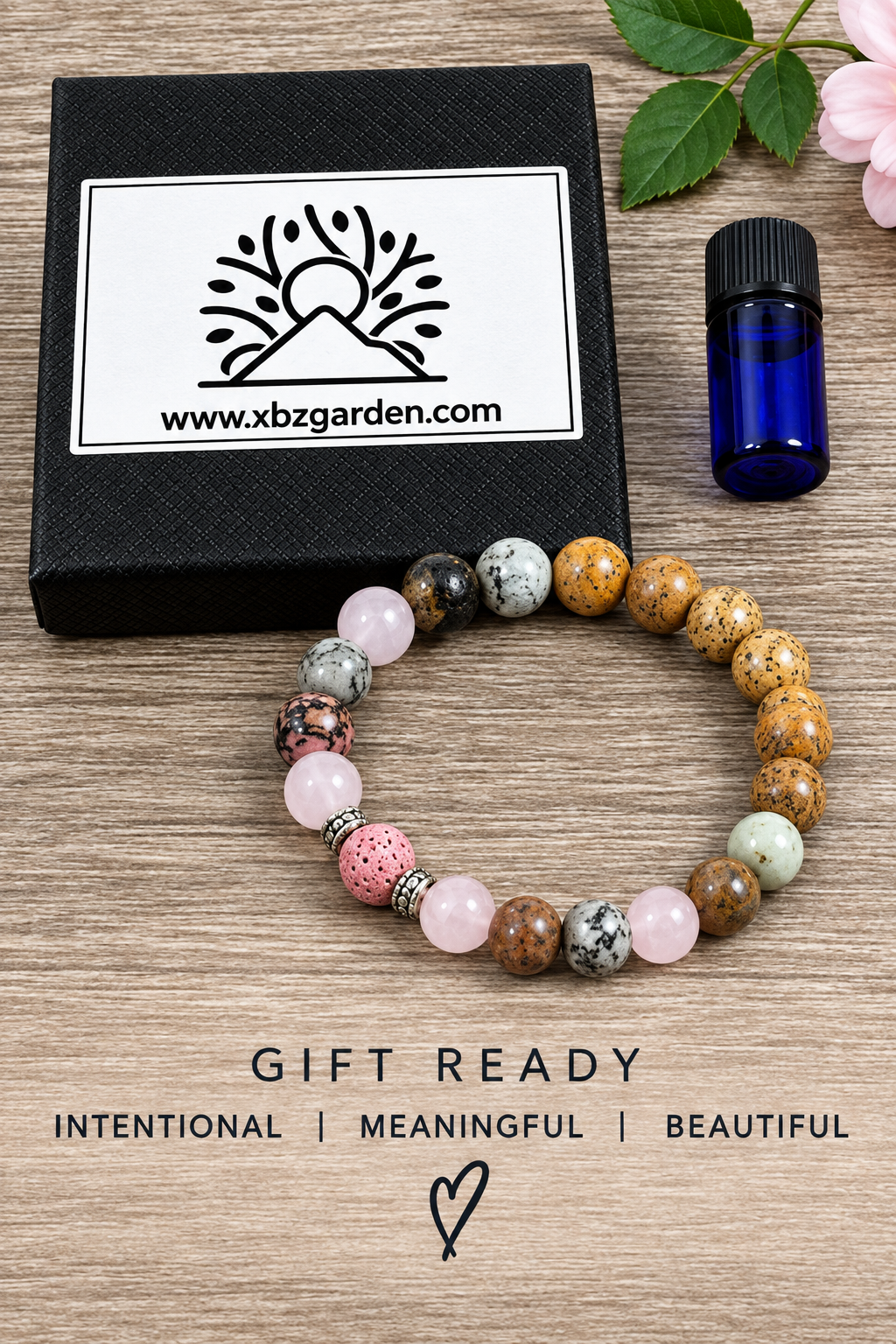 A beaded stretch bracelet made of pink, brown, and gray stones, displayed on a wooden surface next to its black gift box with the brand logo 'xbz garden' and a small blue bottle. The text 'GIFT READY' is printed below.