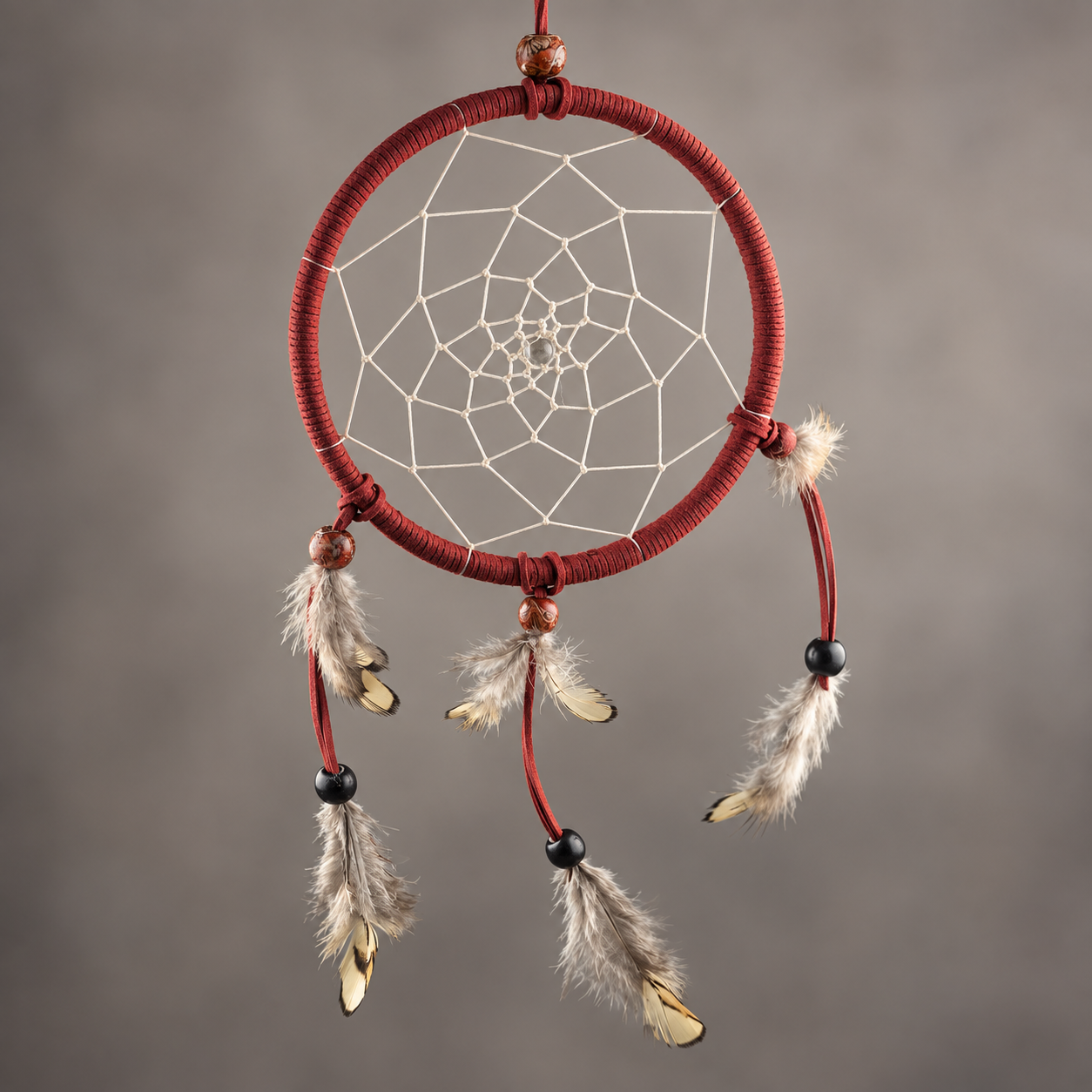 A red dreamcatcher with white webbing, hanging feathers, and wooden beads, centered against a neutral gray background.