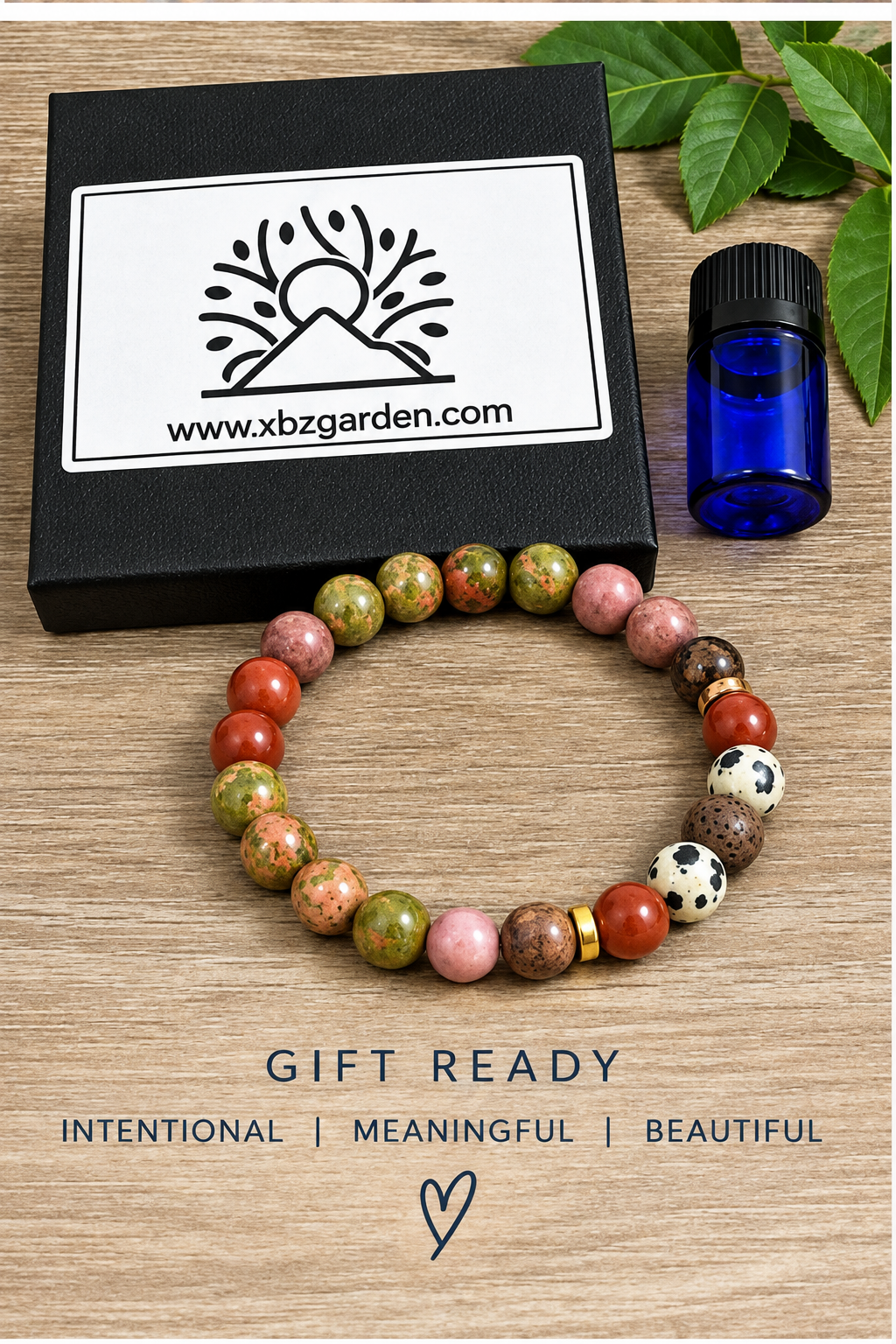 A beaded stretch bracelet made of earthy red, green, and brown stones, displayed on a wooden surface next to a black gift box with the website xbzgarden.com, a small blue bottle, and green leaves.