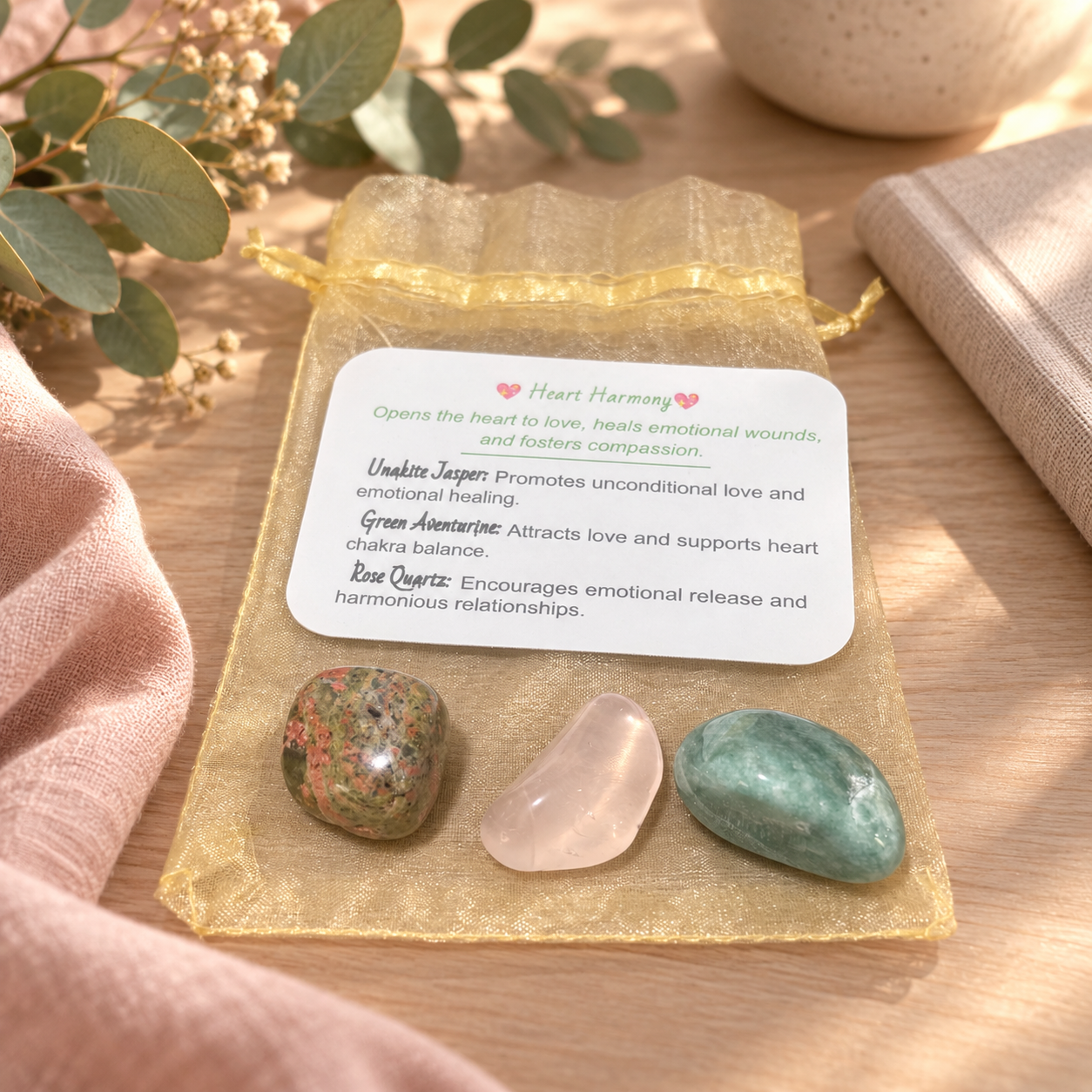 A heart-shaped crystal gift set on a gold drawstring pouch with a white card detailing the benefits of Unakite, Green Aventurine, and Rose Quartz. The set is styled on a wooden surface with a pink fabric, eucalyptus leaves, and a ceramic mug.