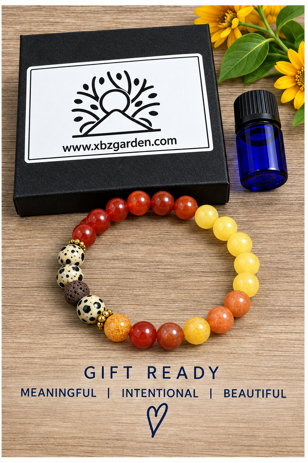 A beaded gemstone bracelet arranged in a circle on a wooden surface, with a black branded gift box, a small blue bottle, and yellow flowers in the background. The text below reads 'GIFT READY' and 'MTENITGFUL | INTENTIONAL | BEAUTIFUL'.