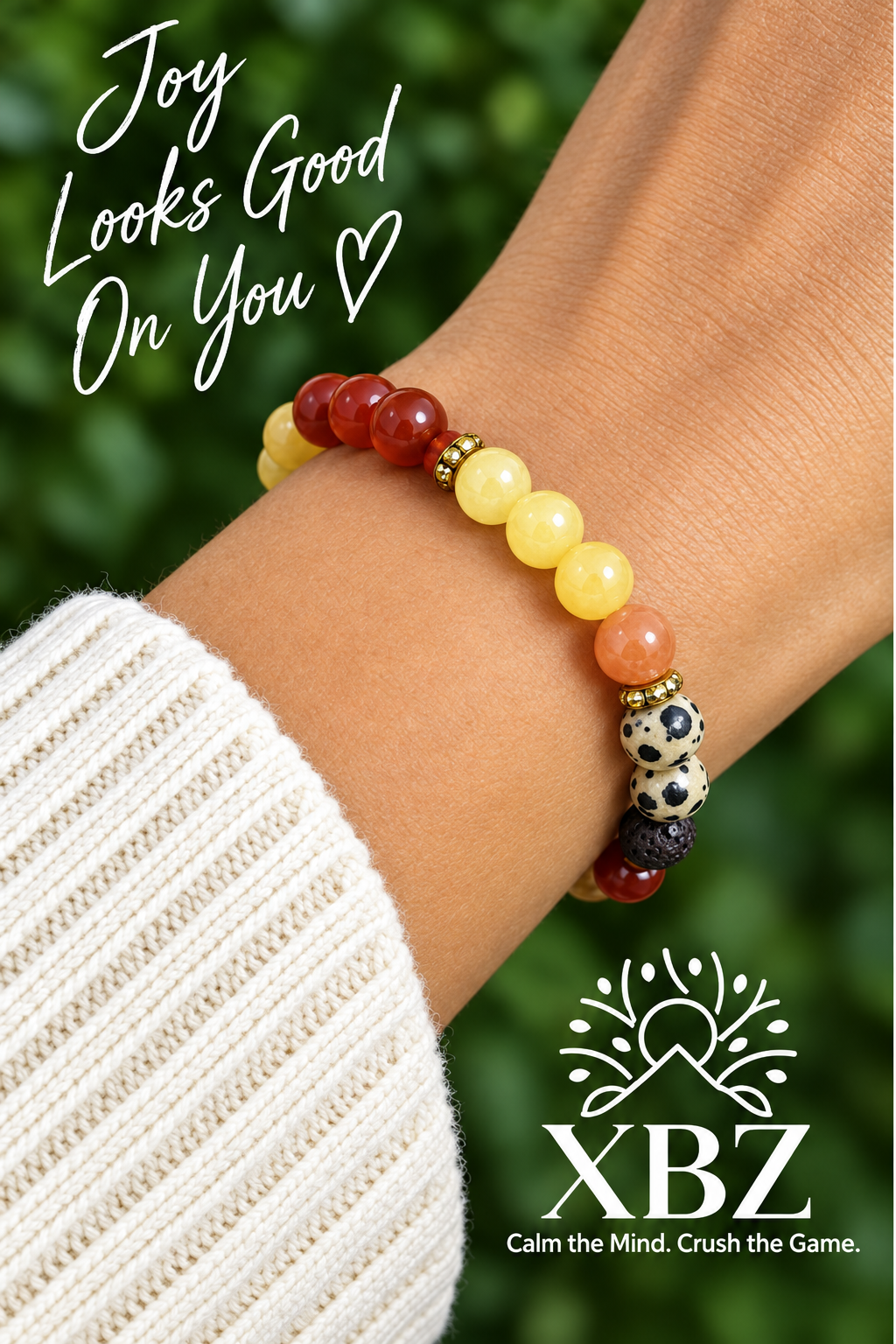 A close-up of a person's wrist wearing a beaded bracelet with red, yellow, and orange beads, alongside a white knit sleeve and green blurred foliage in the background. The text 'Joy Looks Good On You' appears on the left, and the XBZ logo appears at the bottom right.