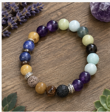 A beaded stretch bracelet made of round gemstone and stone beads in multiple colors, including purple, blue, yellow, green, black, and brown, with decorative metallic spacer beads, displayed on a wooden surface with small plant props.