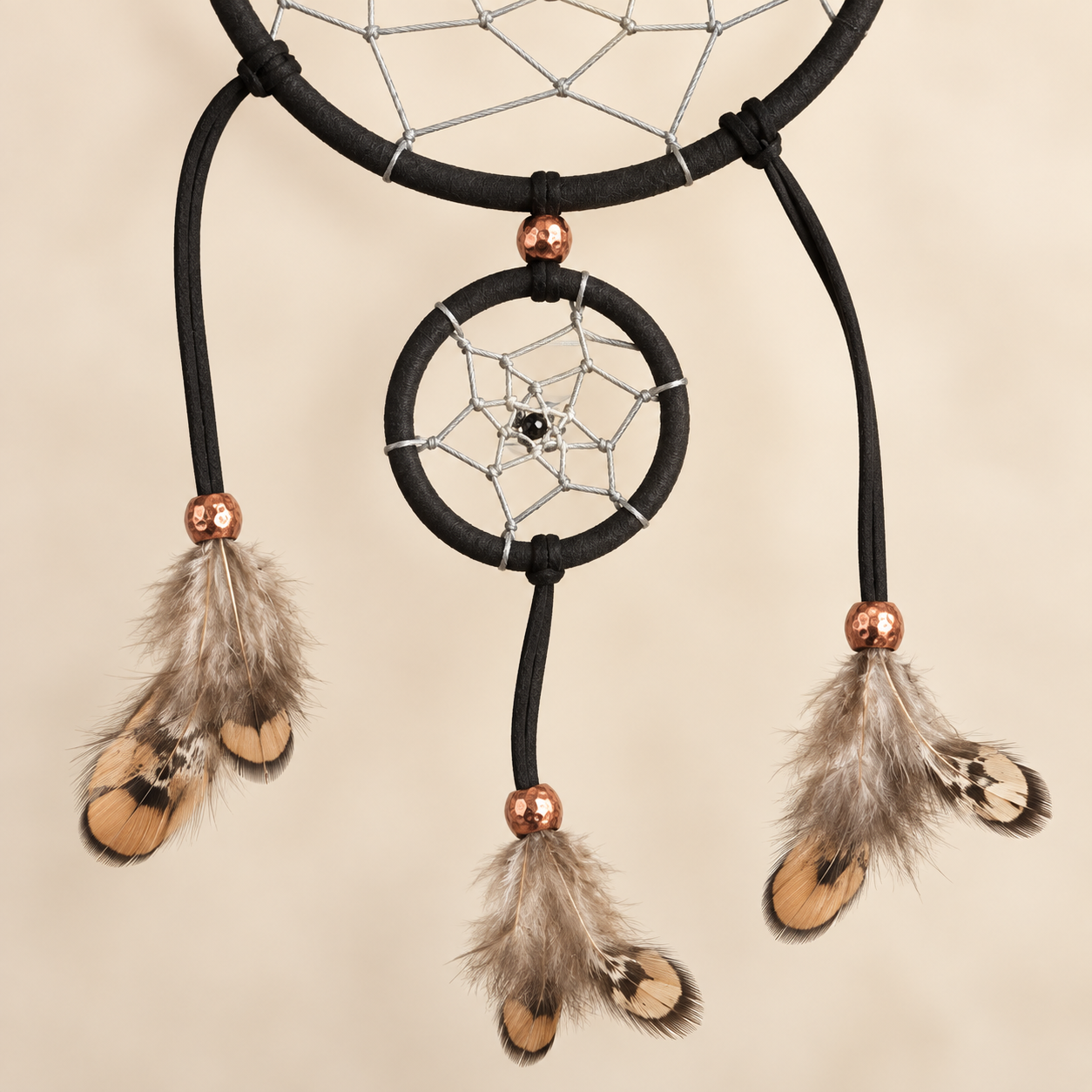 A dreamcatcher with a black wrapped hoop, silver webbing, and three hanging cords with brown and beige feathers, accented by metallic copper beads, shown against a solid beige background.