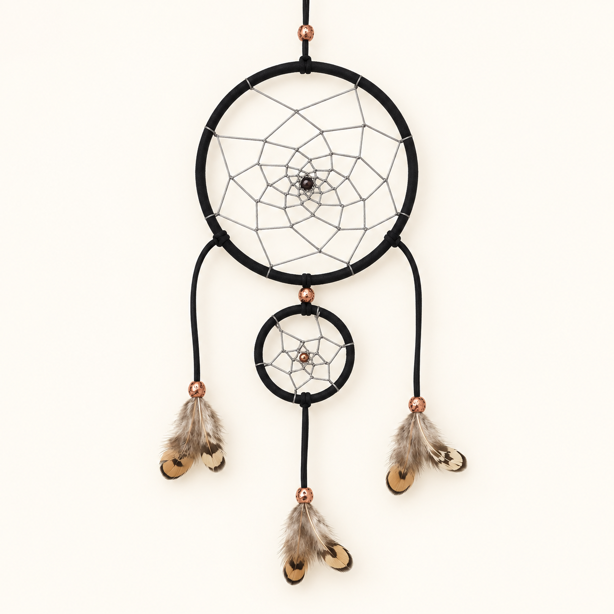 A hanging dreamcatcher wall decor piece with two black hoops, white webbing, small metallic copper beads, and three feather strands in brown, gray, and cream tones, shown against a plain off-white background.