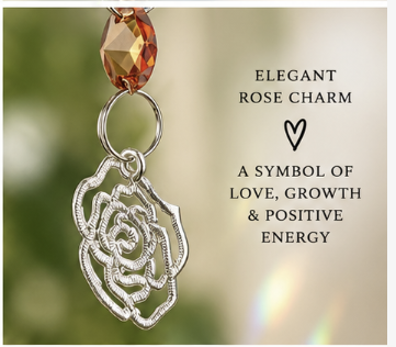 A silver rose charm pendant with a faceted amber-orange bead at the top, shown against a soft blurred green and beige background with text reading 'ELEGANT ROSE CHARM' and 'A SYMBOL OF LOVE, GROWTH & POSITIVE ENERGY'.