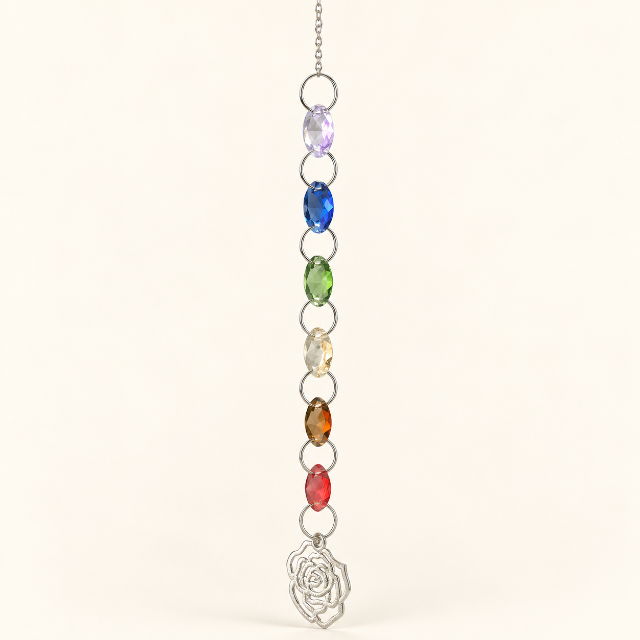 A vertical silver-toned necklace featuring seven colorful oval gemstones in purple, blue, green, clear, amber, red, and rose gold, ending in a silver rose pendant, shown against a plain off-white background.