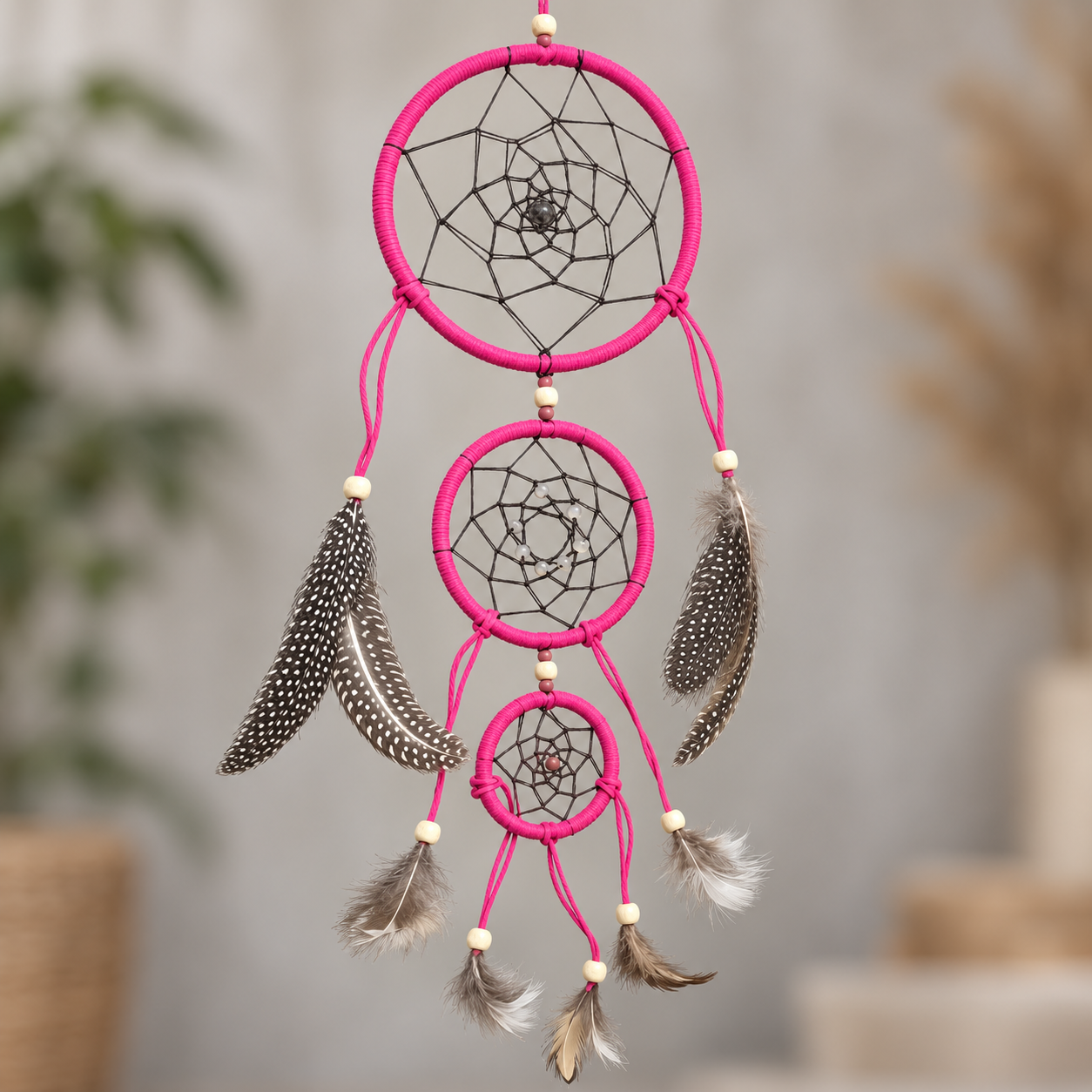 A pink dreamcatcher with three hanging rings, black webbing, and mixed brown, white, and gray feathers, photographed against a softly blurred indoor background.