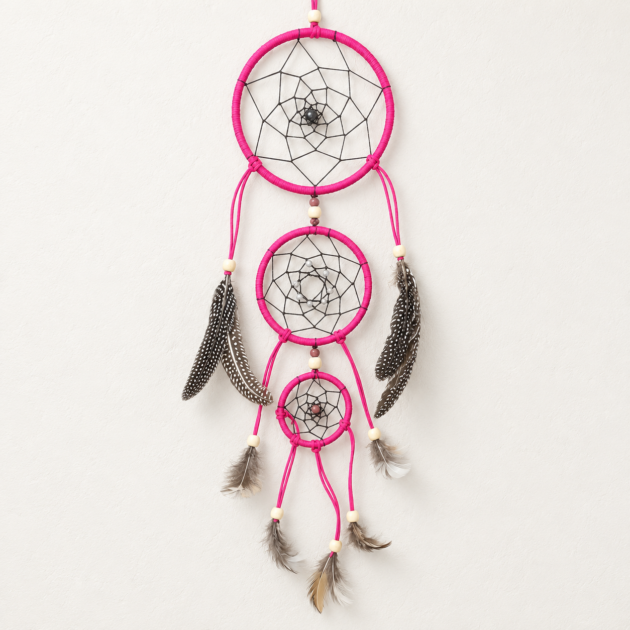 A hanging dreamcatcher with three nested rings in bright pink, black webbing, and dark gray and white feathers, suspended against a plain off-white wall.