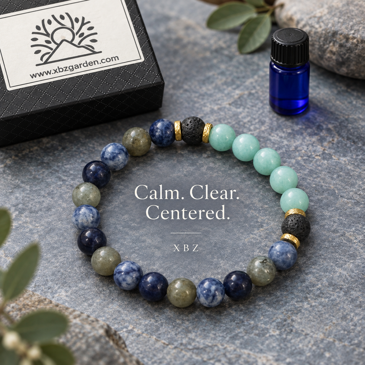 A beaded stretch bracelet arranged in a circle on a gray stone surface, with a small blue essential oil bottle, green leaves, and a black branded box nearby. The bracelet features navy, light blue, mint green, olive green, black, and gold-toned beads.