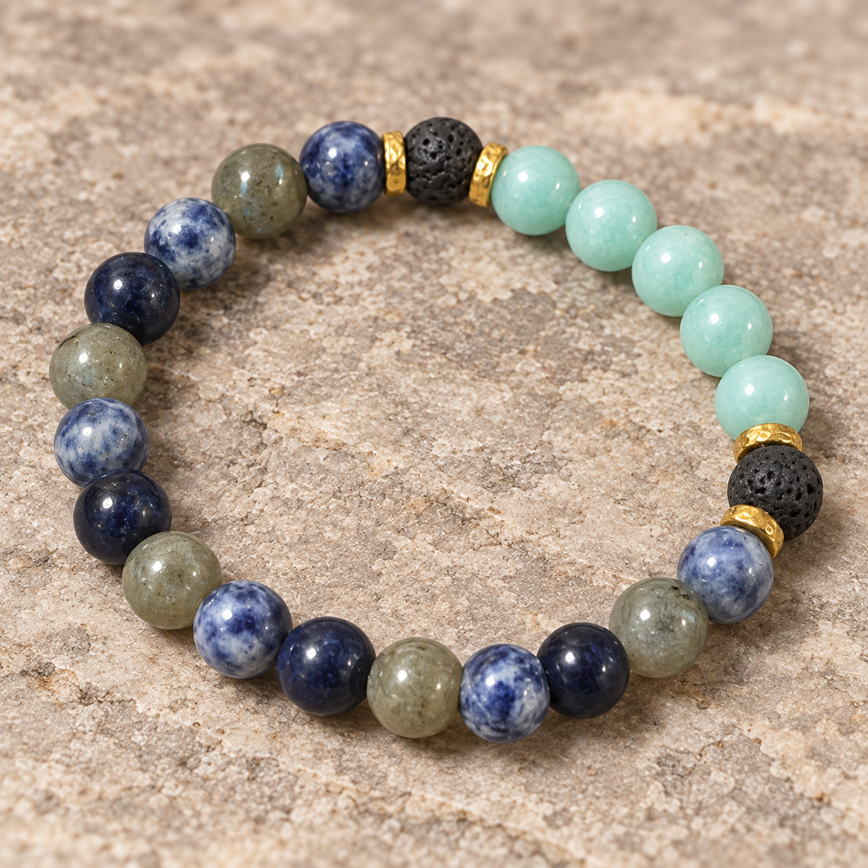 A beaded stretch bracelet arranged in a circle on a textured stone surface. The bracelet features round beads in navy blue, light green, olive green, black lava stone, and gold spacer rings.
