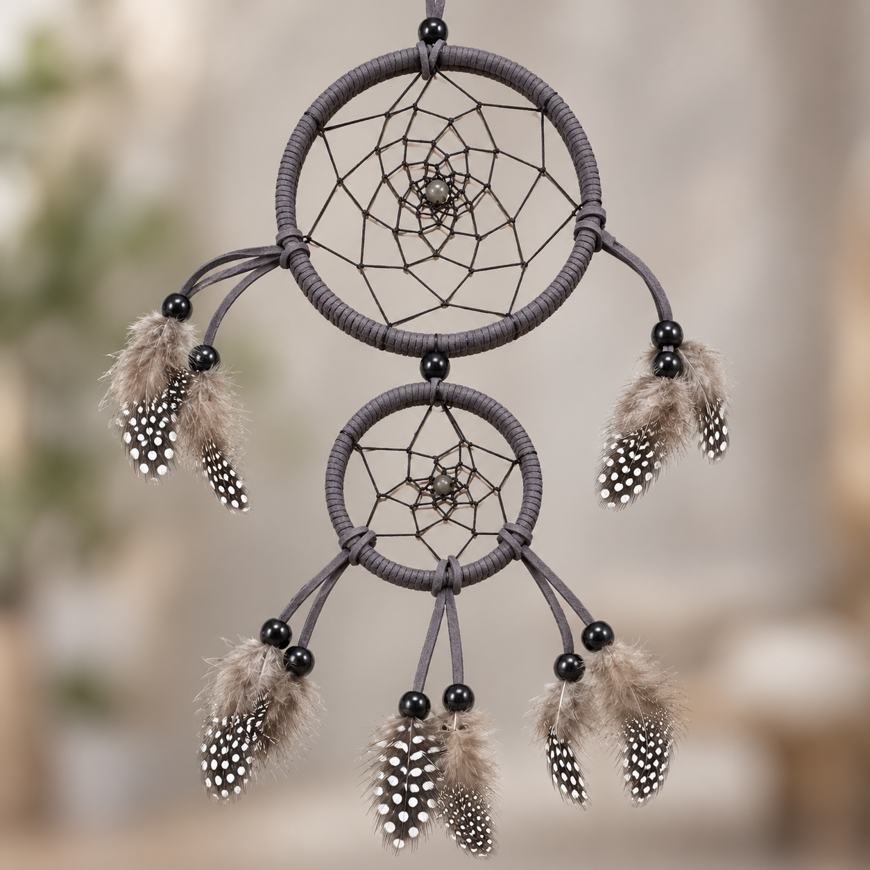 A double-tier dreamcatcher with dark gray wrapped rings, black beads, brown and white feathers, and web patterns, shown against a soft, blurred beige background.