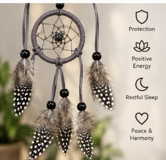 A dark gray wrapped dreamcatcher with black beads and hanging feathers, shown against a softly blurred indoor background with text callouts reading Protection, Positive Energy, Restful Sleep, and Peace & Harmony.