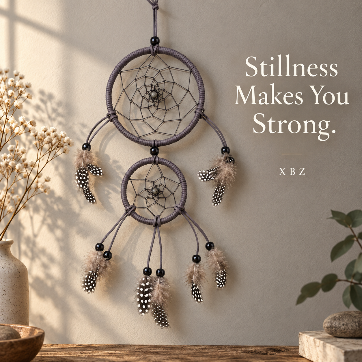 A two-tier dreamcatcher wall hanging with dark gray wrapped rings, black beads, and cream-and-black feathers, displayed against a warm beige wall with dried flowers and plants in the scene. White text on the right reads 'Stillness Makes You Strong.