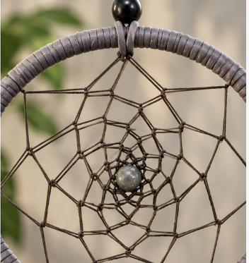 A close-up of a dreamcatcher with a gray wrapped hoop, dark brown webbing, and a central gray stone bead, shown against a softly blurred beige background with hints of green foliage.