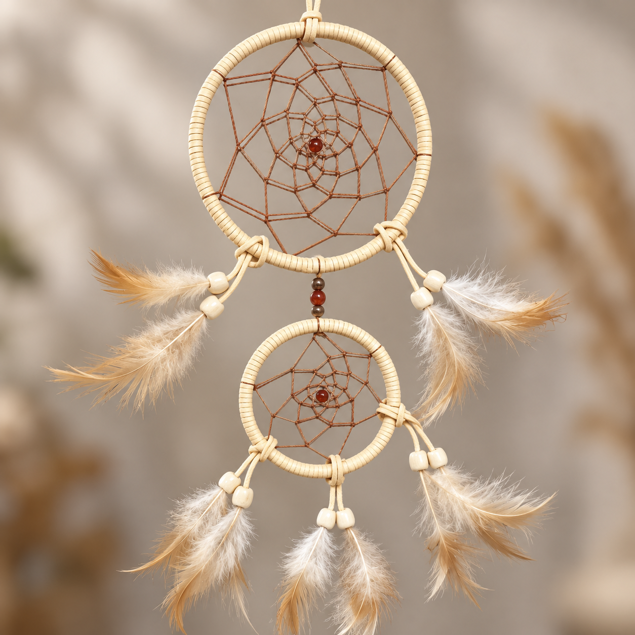 A two-ring dreamcatcher with natural wood wrapped hoops, brown webbing, small white beads, and hanging feathers in cream and tan tones, shown against a softly blurred neutral background.