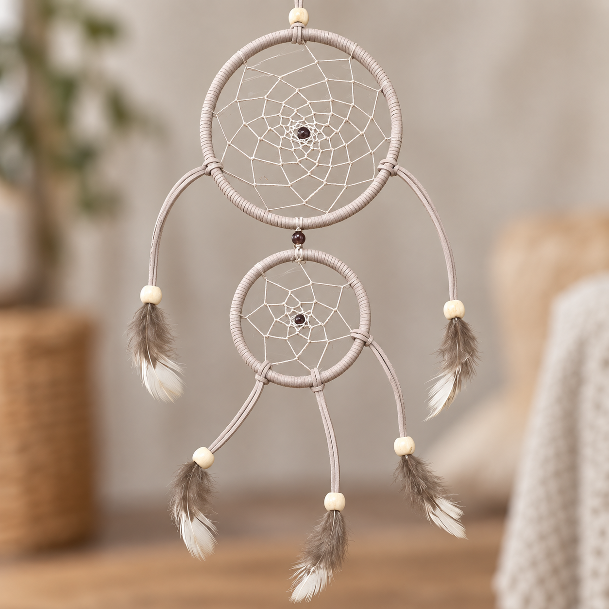 A two-ring dreamcatcher with beige wrapped cords, white and brown feathers, and small cream beads, hanging against a softly blurred indoor background.