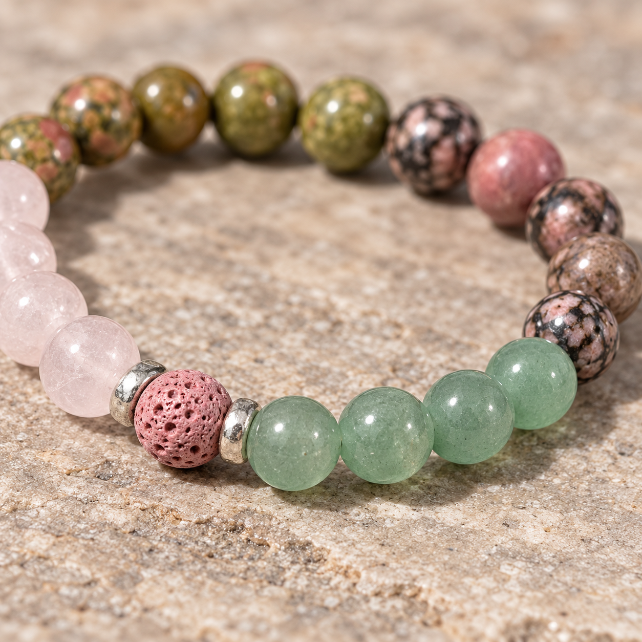 A beaded stretch bracelet displayed on a textured stone surface. The bracelet features a mix of green, pink, brown, and pale pink beads, including polished round beads, mottled stone beads, a pink lava stone, and silver spacer rings.