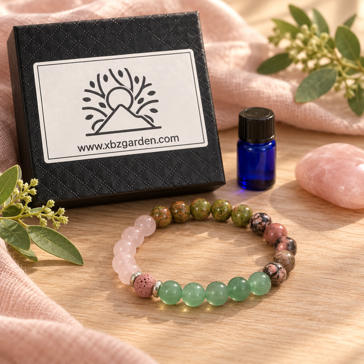 A beaded stretch bracelet with light pink, green, and mottled brown beads is displayed on a wooden surface next to a black branded gift box, a small blue essential oil bottle, and soft pink fabric with green leaves.