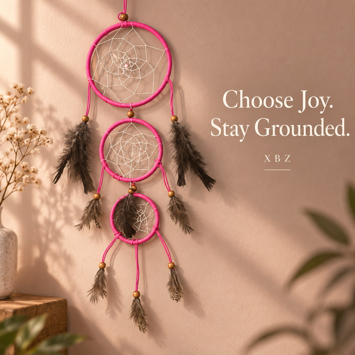 A pink dreamcatcher wall hanging with three nested rings, white webbing, and dark brown feathers, displayed on a beige wall with decorative dried flowers and plants. Text on the right reads 'Choose Joy.