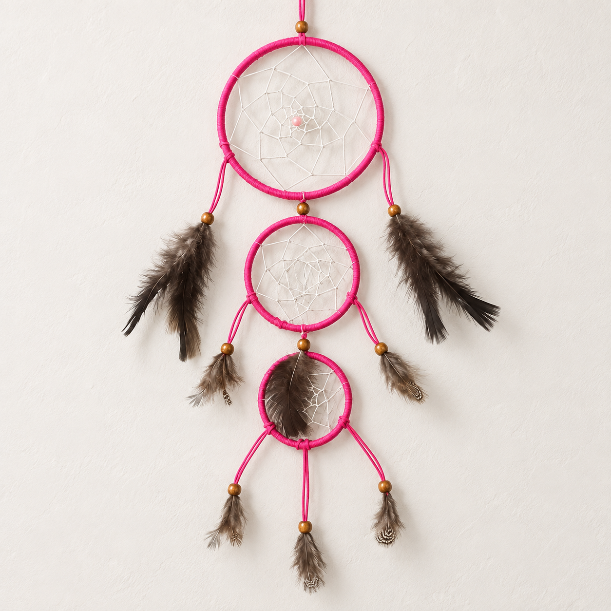 A three-tier dreamcatcher with bright pink wrapped rings, white webbing, brown feathers, and small gold beads, hanging against a textured off-white wall.