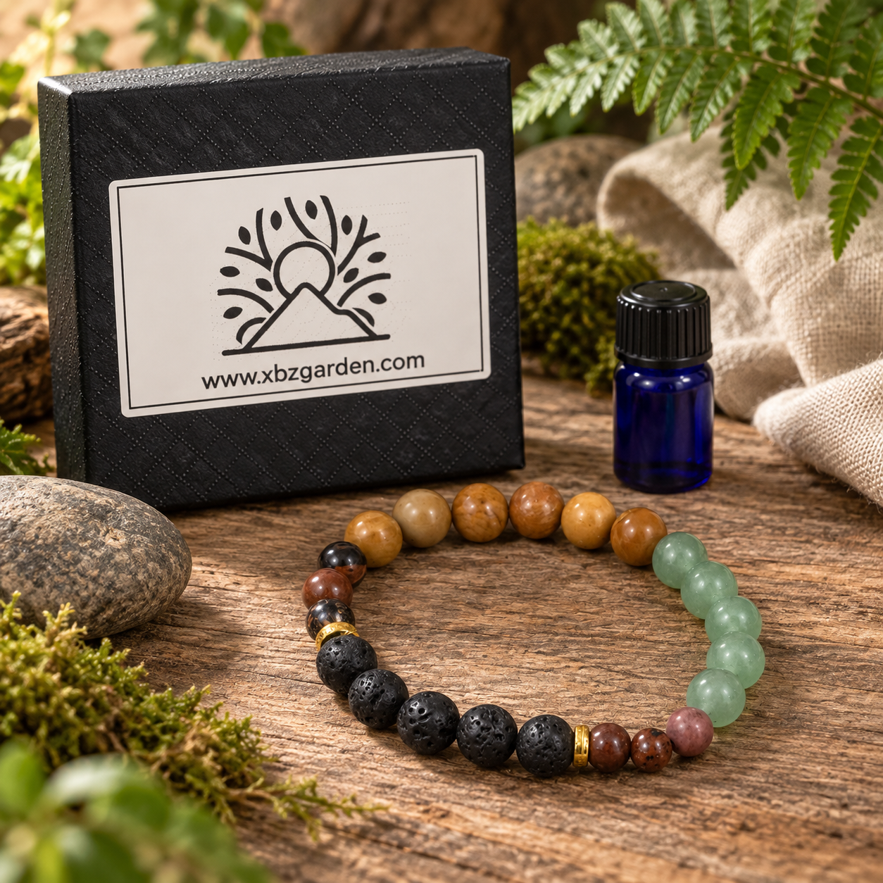 A beaded bracelet with black lava stones, brown and tan wood beads, a light green segment, and small pink and gold accent beads, displayed on a rustic wooden surface with moss, a small blue bottle, and a black branded box reading xbzgarden.com.