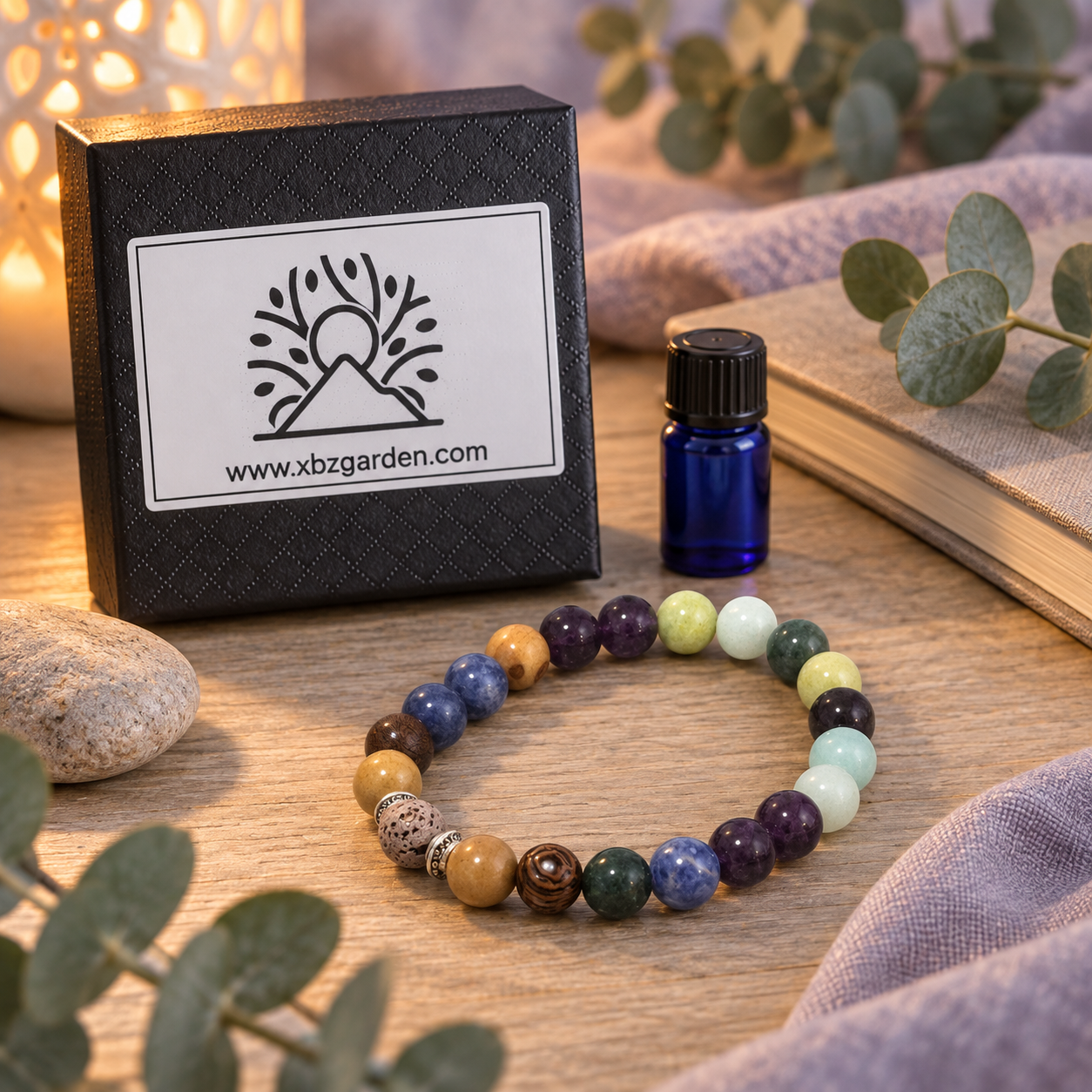 A multicolor beaded bracelet made of polished stone beads is displayed on a wooden surface next to its black branded gift box and a small cobalt blue essential oil bottle. The scene is styled with eucalyptus leaves, a book, and a soft purple cloth.