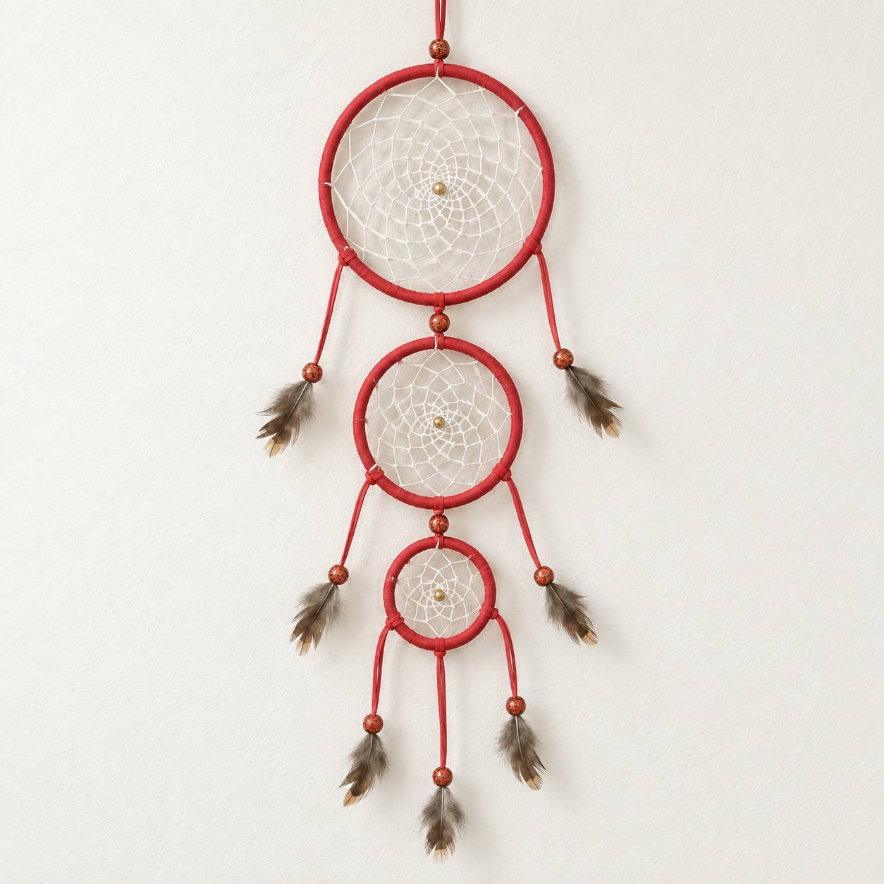 A red three-ring dreamcatcher with white webbing, brown beads, and hanging brown feathers, shown against a plain off-white wall.