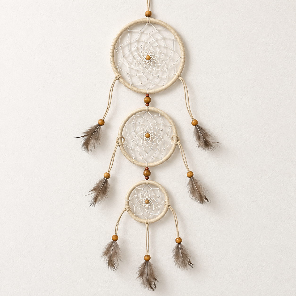 A three-tier dreamcatcher wall hanging with natural beige woven hoops, white webbing, small brown wooden beads, and hanging brown feathers, shown against a plain off-white wall.