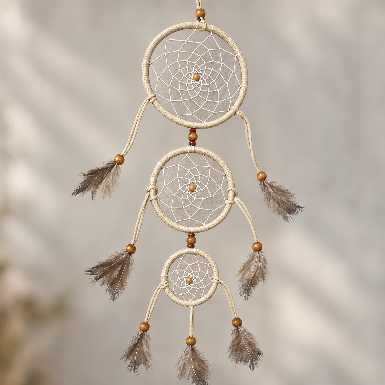A three-tier dreamcatcher wall hanging with cream-colored wrapped rings, white webbing, small wooden beads, and brown feathers, shown against a softly blurred neutral background.