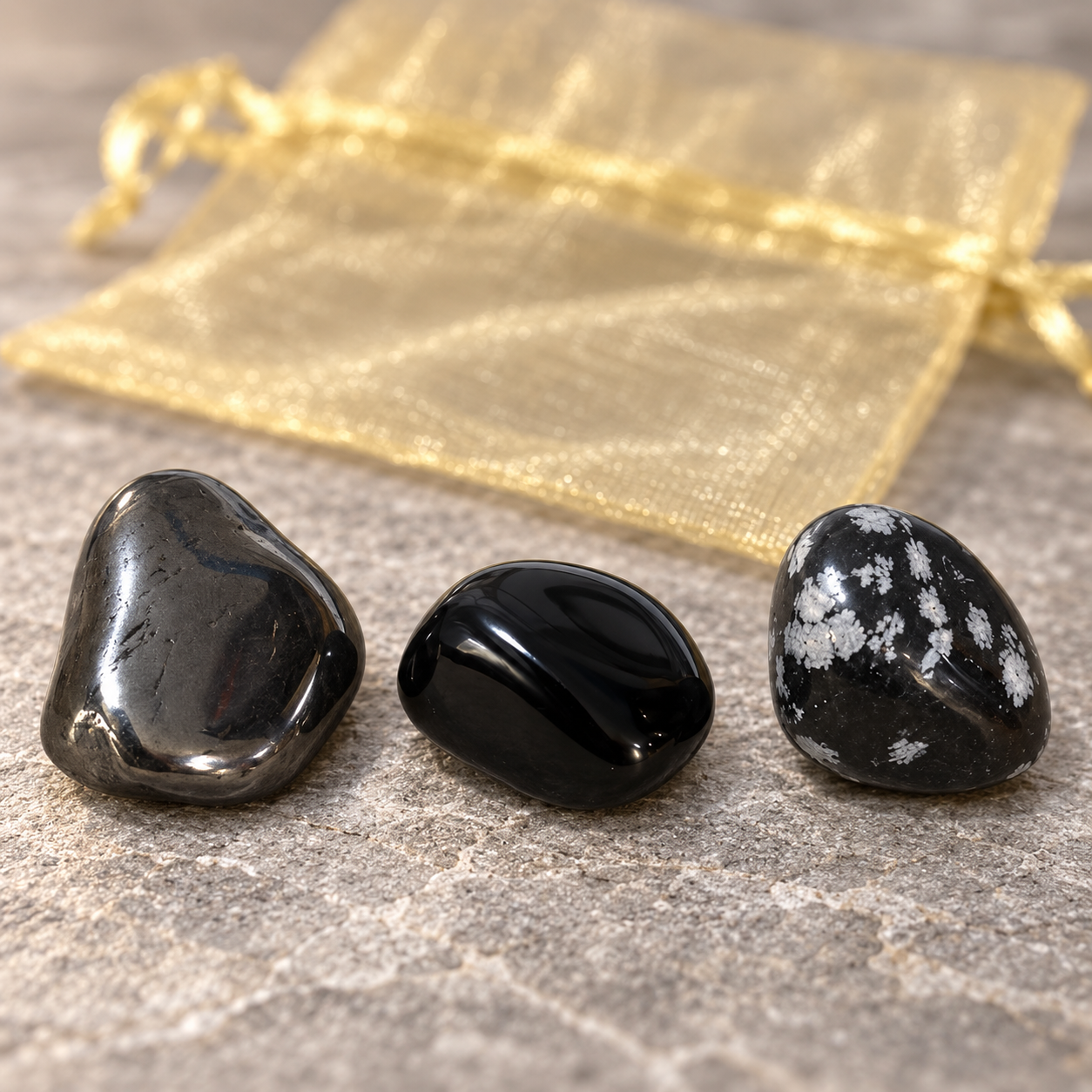 Three polished black tumbled stones with white speckles sit on a gray stone surface, with a sheer gold drawstring pouch blurred in the background.