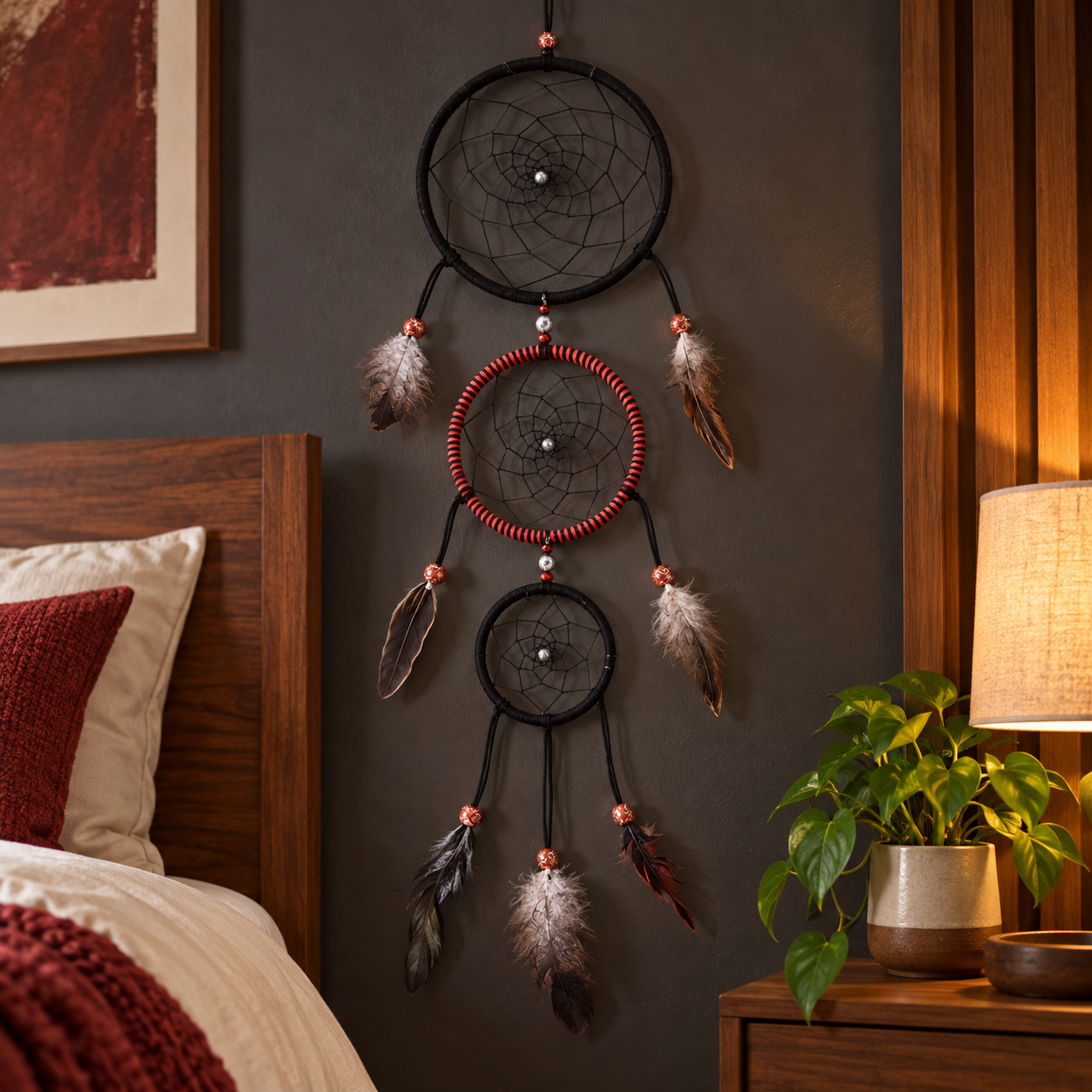 A large wall-mounted dreamcatcher with three nested circular hoops hangs on a dark gray wall in a bedroom setting, with a bed, lamp, and plant visible nearby.