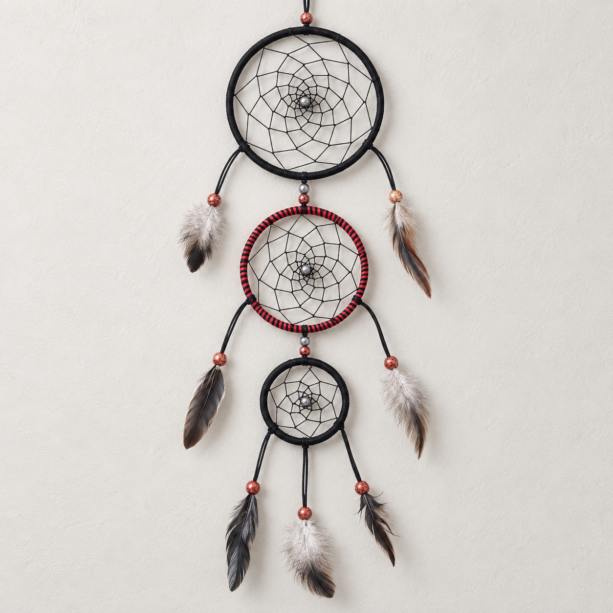 A three-tier dreamcatcher wall hanging with black wrapped hoops, red accent beads, white and brown feathers, and copper-colored beads, hanging against a textured off-white wall.