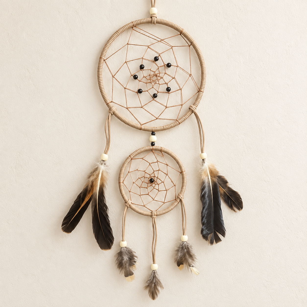 A double-ring dreamcatcher wall hanging with natural tan wrapped rings, brown webbing, small black beads, and hanging feathers in black, brown, and gray on a light beige textured wall.